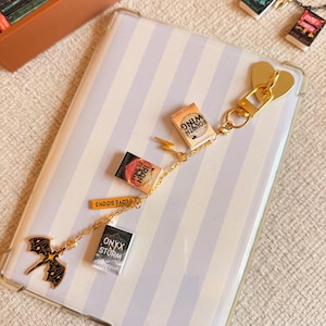 May include: A gold-colored keychain with miniature book charms, including titles like "Fourth Wing" and "Onyx Storm." The keychain features a heart-shaped clasp and a dragon charm. The background is a striped phone case.