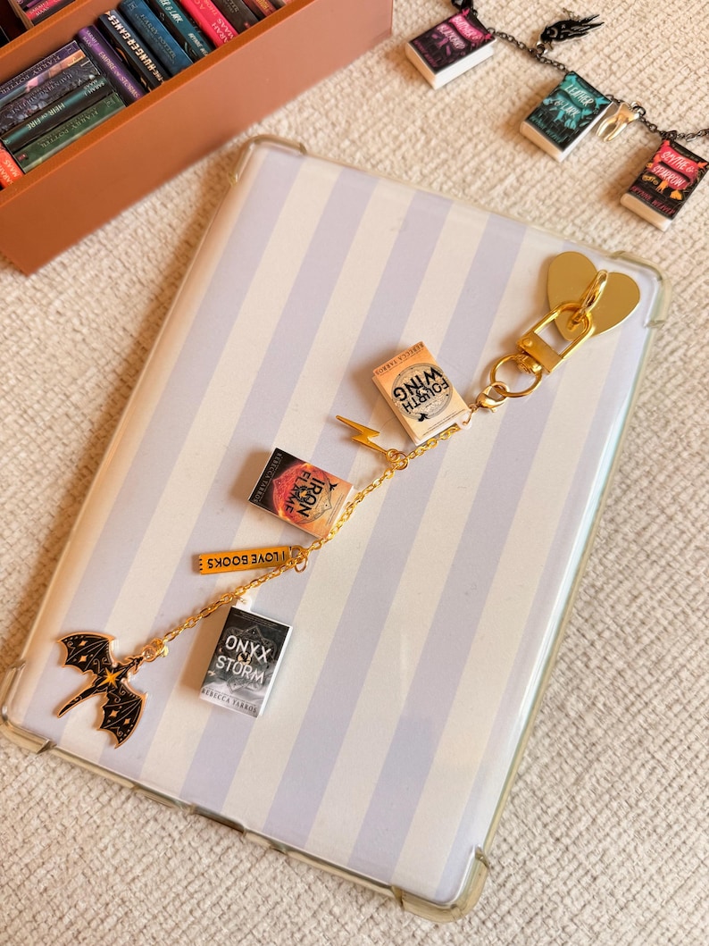 May include: A gold-colored keychain with miniature book charms, including titles like "Fourth Wing" and "Onyx Storm." The keychain features a heart-shaped clasp and a dragon charm. The background is a striped phone case.