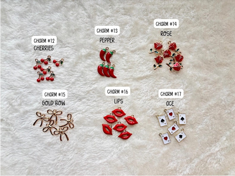 May include: A collection of charms with various designs, including cherries, peppers, roses, gold bows, red lips, and playing card aces. Each charm is labeled with a number and a descriptive word. The charms are arranged on a white, textured surface.