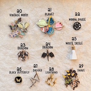 May include: A collection of fantasy-themed charms, including a vintage moth, planets, a moon phase, dragons, a white tassel, a black butterfly, daggers, lightning bolts, and a snake. The charms are in various colors and designs.