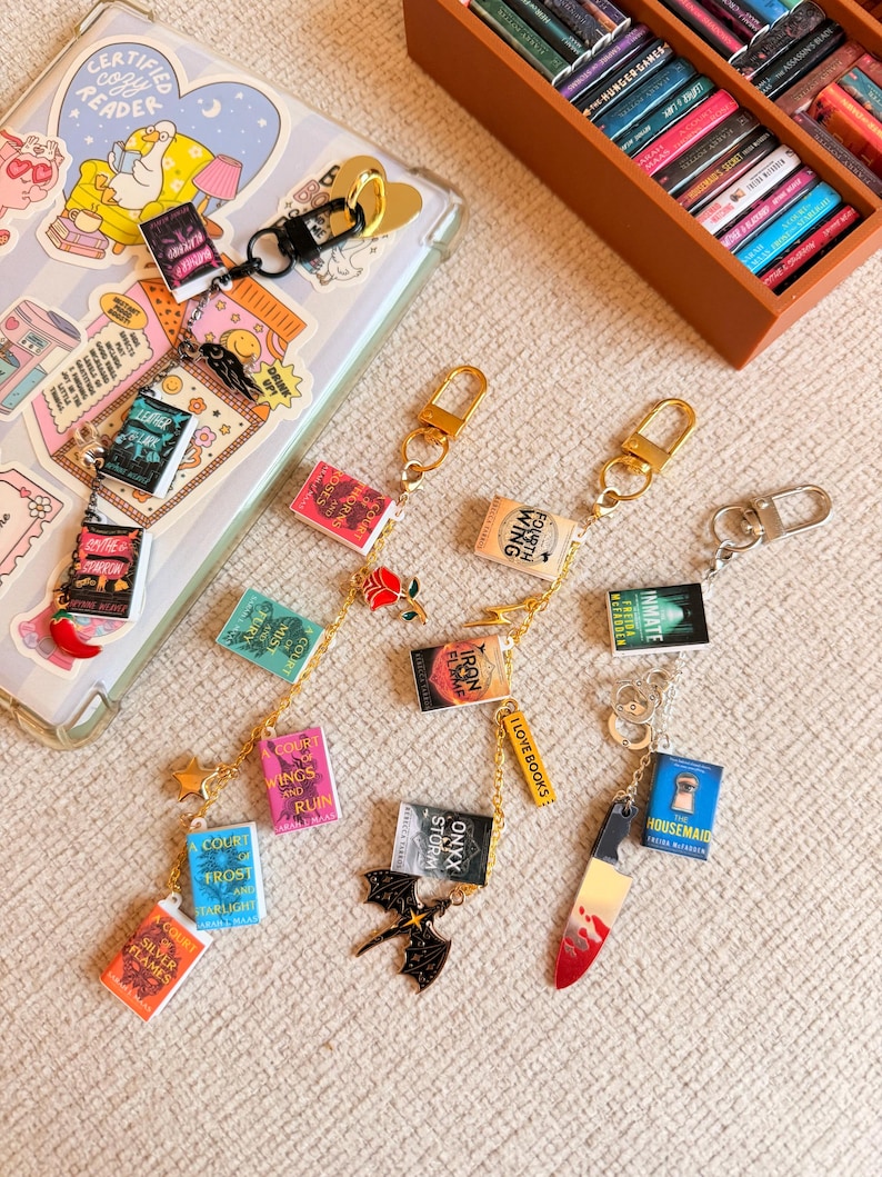 May include: Miniature book charms on keychains. The charms feature book titles like "Court of Silver Flames" and "Housemaid." Gold-colored chains connect the books to the key rings. A decorative knife charm and a black dragon charm are also included.