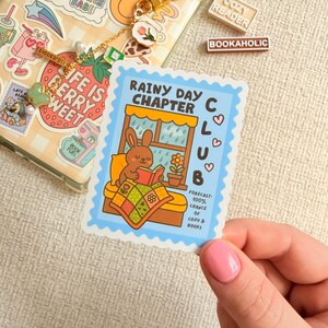 May include: A sticker with a blue border, depicting a cartoon bunny reading a book by a window. Text on the sticker reads "Rainy Day Chapter Club" and "Forecast: 100% chance of cozy & books."