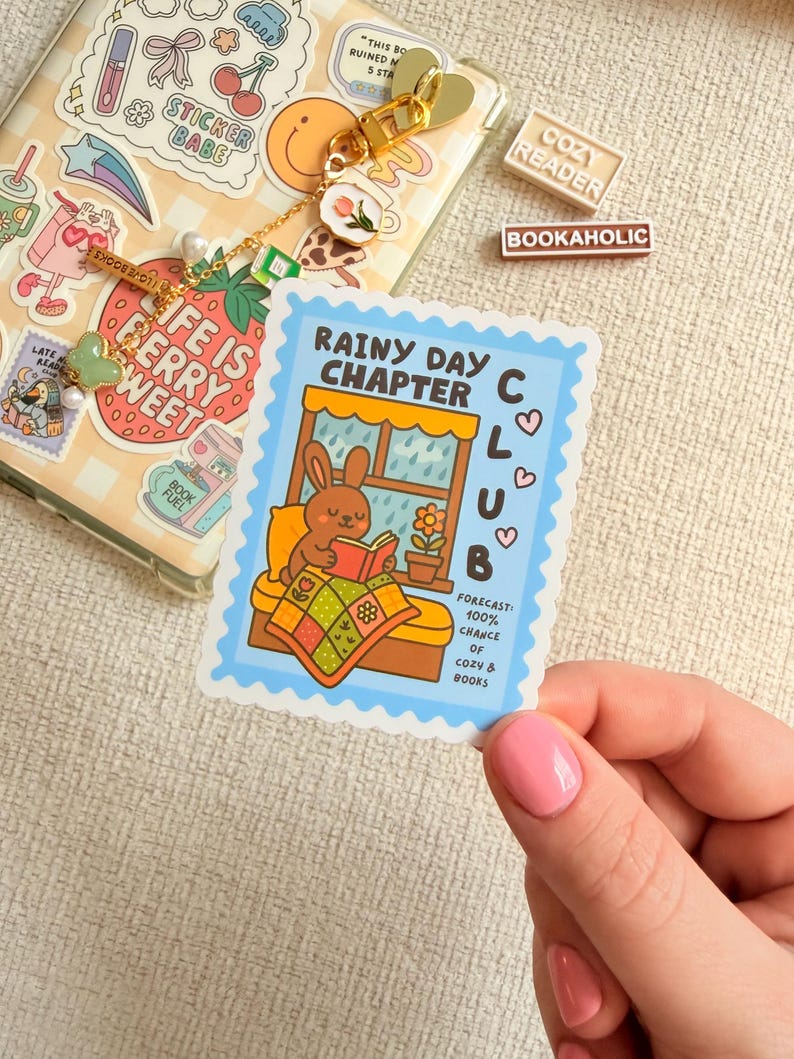 May include: A sticker with a blue border, depicting a cartoon bunny reading a book by a window. Text on the sticker reads "Rainy Day Chapter Club" and "Forecast: 100% chance of cozy & books."