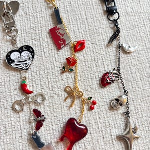 May include: A collection of keychains with various charms. Charms include a skeleton couple in a heart, a red chili pepper, handcuffs, a butterfly, a cleaver, a rose, lips, a bow, cherries, a dripping heart, a skull, a star, a raven, and a crescent moon.