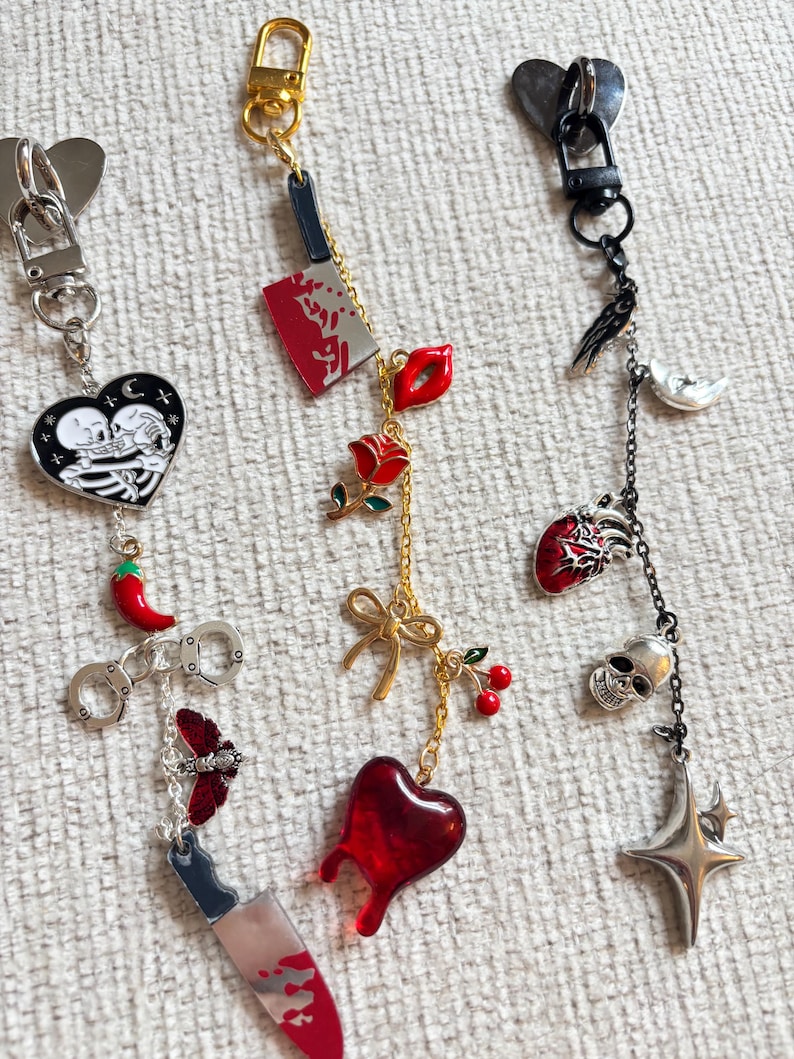 May include: A collection of keychains with various charms. Charms include a skeleton couple in a heart, a red chili pepper, handcuffs, a butterfly, a cleaver, a rose, lips, a bow, cherries, a dripping heart, a skull, a star, a raven, and a crescent moon.
