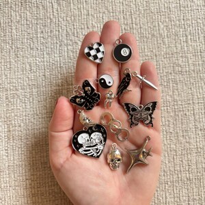 May include: A collection of silver and enamel charms in various shapes, including a butterfly, skull, heart, and an eight ball. The charms are in black, white, and silver tones, with intricate details.