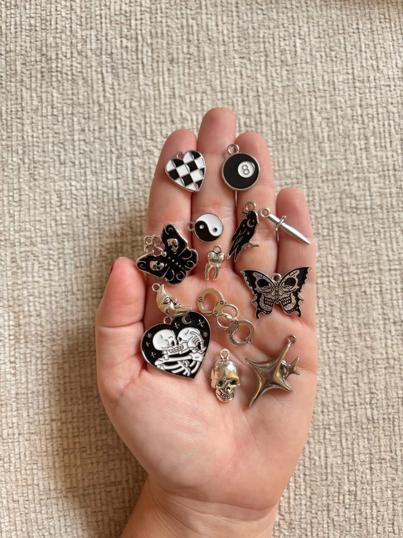 May include: A collection of silver and enamel charms in various shapes, including a butterfly, skull, heart, and an eight ball. The charms are in black, white, and silver tones, with intricate details.