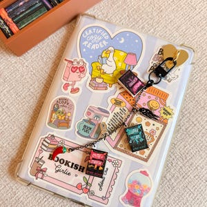 May include: A clear phone case decorated with stickers, including a heart-shaped "Certified Cozy Reader" design. A keychain with miniature book charms hangs from the case. The case is on a striped background, with a small wooden bookshelf in the upper left corner.