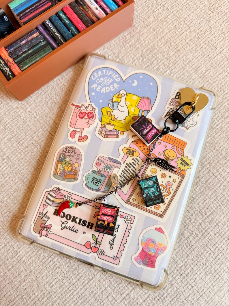 May include: A clear phone case decorated with stickers, including a heart-shaped "Certified Cozy Reader" design. A keychain with miniature book charms hangs from the case. The case is on a striped background, with a small wooden bookshelf in the upper left corner.