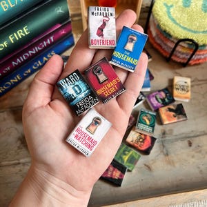 May include: Miniature books with titles like "The Boyfriend" and "The Housemaid" are held in a hand. The books have colorful covers and the author's name, Freida McFadden, is visible. Other books are stacked in the background.