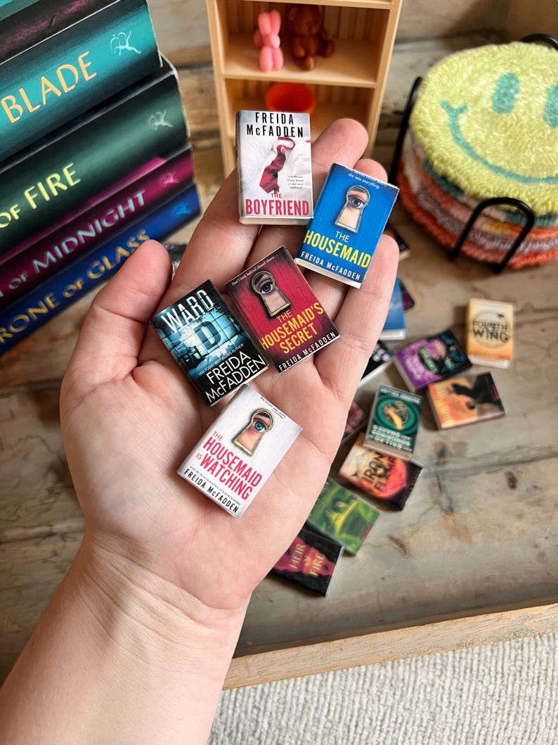 May include: Miniature books with titles like "The Boyfriend" and "The Housemaid" are held in a hand. The books have colorful covers and the author's name, Freida McFadden, is visible. Other books are stacked in the background.