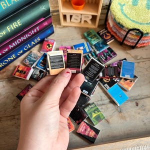 May include: A collection of miniature books, some stacked, others scattered, with titles like "Blade" and "Heir of Fire." A small wooden shelf with the letters "TBR" and a smiley face coaster are also visible. The books are in various colors.