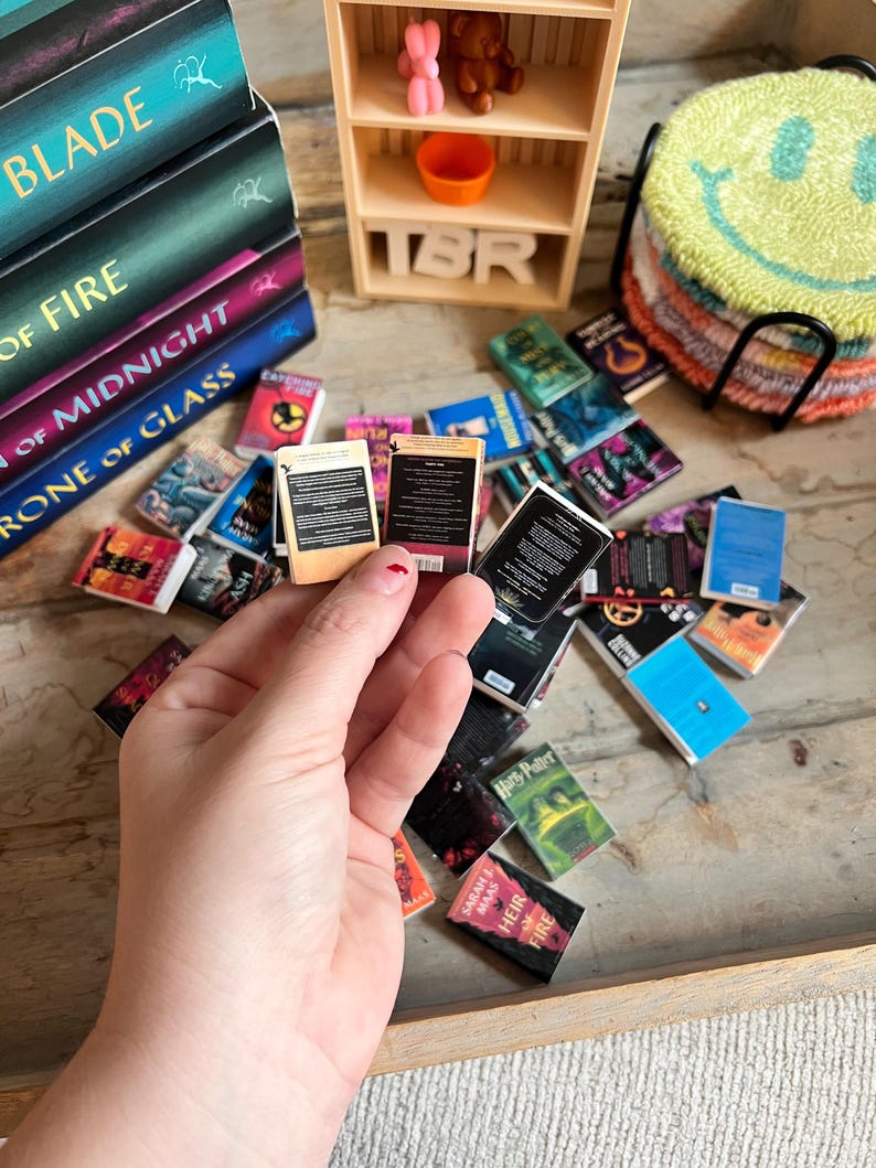 May include: A collection of miniature books, some stacked, others scattered, with titles like "Blade" and "Heir of Fire." A small wooden shelf with the letters "TBR" and a smiley face coaster are also visible. The books are in various colors.