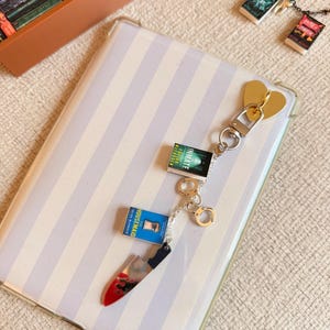 May include: A gold heart-shaped keychain with a silver clasp, featuring miniature book charms and a knife charm with a red blood splatter design. The keychain is on a striped background.