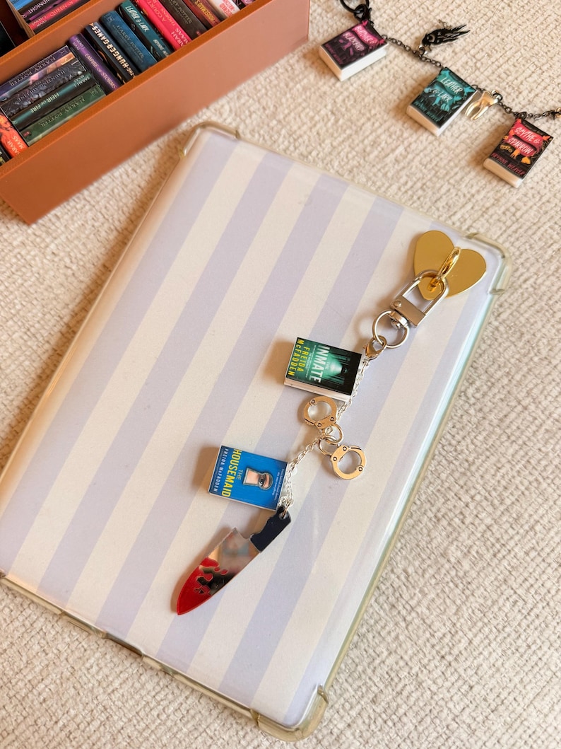 May include: A gold heart-shaped keychain with a silver clasp, featuring miniature book charms and a knife charm with a red blood splatter design. The keychain is on a striped background.