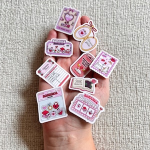 May include: A collection of pink and white romance-themed stickers. The stickers feature hearts, books, and the words "Romance Reader Soda" and "Romance Book Club". The stickers are held in a hand.