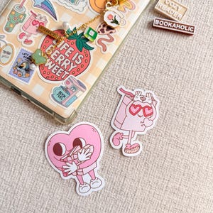 May include: A phone case covered in stickers, a gold keychain with charms, and several stickers. The phone case has a strawberry sticker with the text "LIFE IS BERRY WEET". Two rectangular stickers read "COZY READER" and "BOOKAHOLIC".