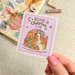 May include: A pink sticker with a scalloped edge shows a cartoon duck reading in bed under a crescent moon and stars. The sticker reads "Bedtime Chapter Club" and "A page a night keeps the worries light."