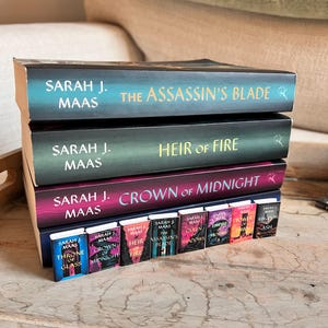 May include: A stack of books by Sarah J. Maas, including "The Assassin's Blade," "Heir of Fire," and "Crown of Midnight." A row of miniature books with the same titles are in front. The books have colorful covers with titles in gold and white.