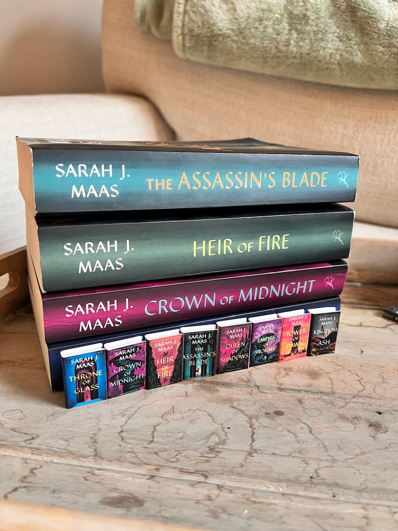 May include: A stack of books by Sarah J. Maas, including "The Assassin's Blade," "Heir of Fire," and "Crown of Midnight." A row of miniature books with the same titles are in front. The books have colorful covers with titles in gold and white.