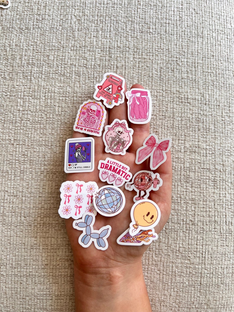 May include: A collection of pink and white stickers featuring various designs, including a skeleton, a disco ball, a balloon, a bow, and a jar of pickles. The stickers are arranged on a hand.