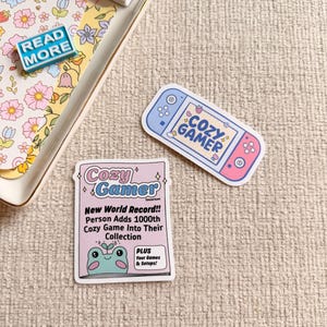 May include: A selection of stickers and miniature books. The stickers feature a pink and blue game console with "COZY GAMER" text, and a pink sticker with a frog graphic and text that reads "New World Record!! Person Adds 1000th Cozy Game Into Their Collection".