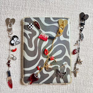 May include: A clear phone case with a black and white zebra print design, adorned with various charms. Charms include a heart, a coffin, a knife, a butterfly, and a skull. The charms are attached to the case with gold and silver chains.