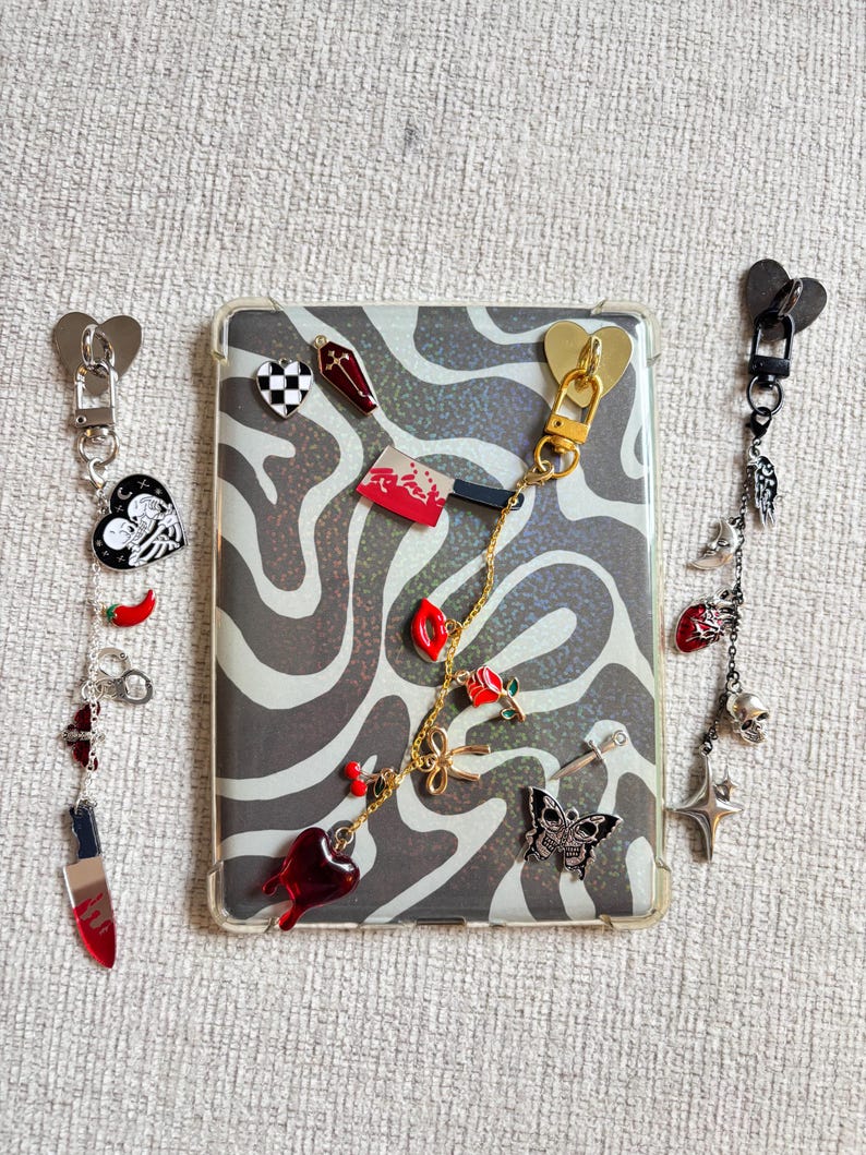 May include: A clear phone case with a black and white zebra print design, adorned with various charms. Charms include a heart, a coffin, a knife, a butterfly, and a skull. The charms are attached to the case with gold and silver chains.