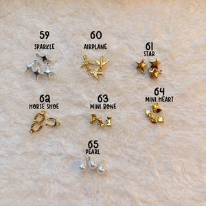 May include: An assortment of gold and silver charms, including stars, airplanes, hearts, and pearls, are displayed on a soft, white surface. The image includes text labels such as "SPARKLE," "AIRPLANE," and "PEARL." The text "GOLD & GIRLY CHARMS" is also visible.