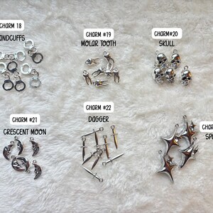 May include: A collection of silver-colored charms, including handcuffs, molar teeth, skulls, crescent moons, daggers, and starbursts. Each charm is labeled with a number and description. The charms are displayed on a white, textured surface.