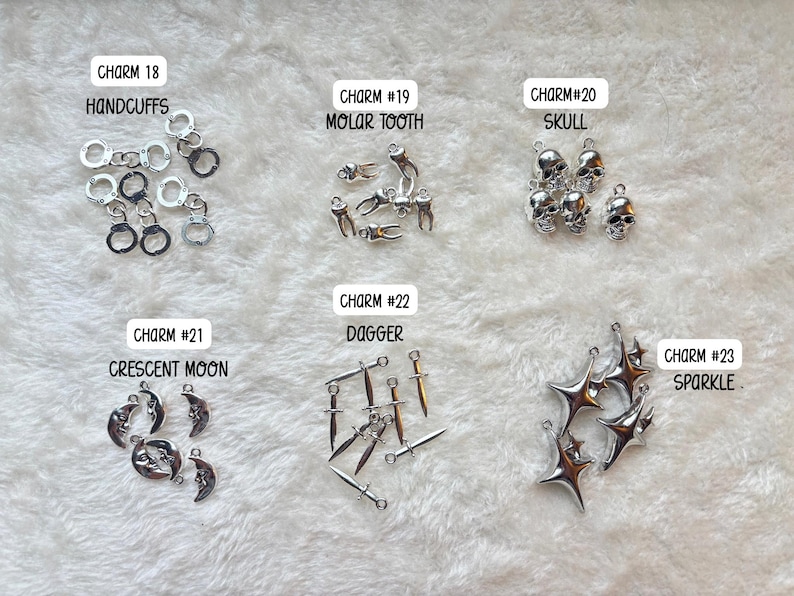 May include: A collection of silver-colored charms, including handcuffs, molar teeth, skulls, crescent moons, daggers, and starbursts. Each charm is labeled with a number and description. The charms are displayed on a white, textured surface.