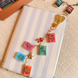 May include: A gold-colored keychain with miniature book charms. The charms feature book titles in various colors, including pink, blue, green, and orange. A gold heart-shaped clasp and a star charm are also present. The background includes a striped phone case and a box of miniature books.