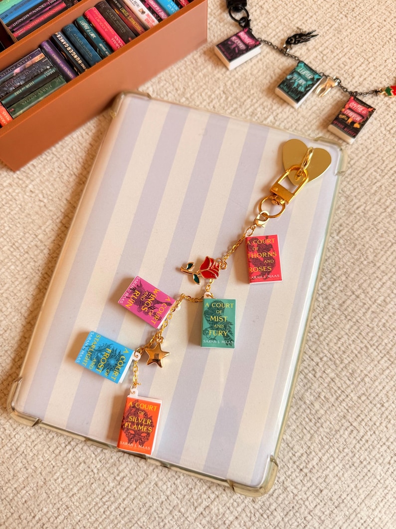 May include: A gold-colored keychain with miniature book charms. The charms feature book titles in various colors, including pink, blue, green, and orange. A gold heart-shaped clasp and a star charm are also present. The background includes a striped phone case and a box of miniature books.