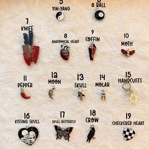 May include: A collection of dark romance charms, including a yin-yang symbol, an 8-ball, a knife, an anatomical heart, a coffin, a moth, a pepper, a moon, a skull, a molar, handcuffs, a kissing skull, a skull butterfly, a crow, and a checkered heart.