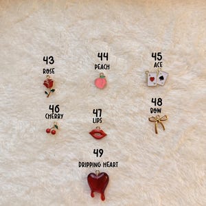 May include: A collection of colorful charms, including a red rose, a peach, playing cards, cherries, red lips, a gold bow, and a dripping heart. The charms are numbered and displayed on a textured white surface. The text "SMUTTY CHARMS" is visible.
