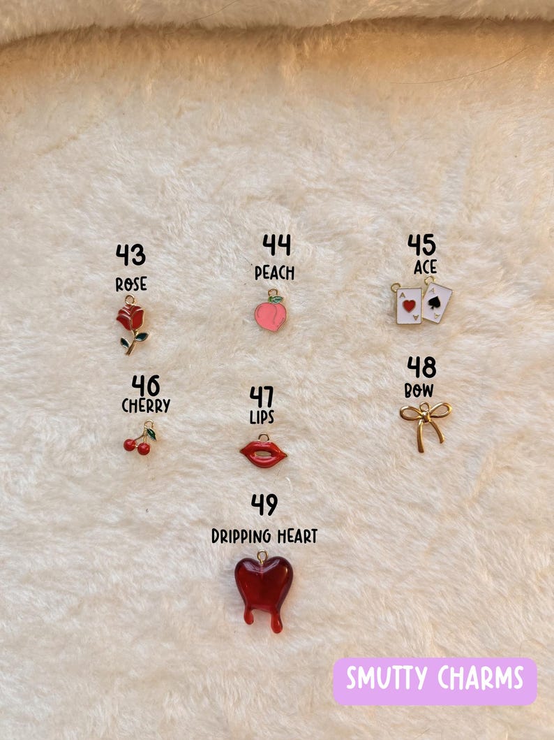 May include: A collection of colorful charms, including a red rose, a peach, playing cards, cherries, red lips, a gold bow, and a dripping heart. The charms are numbered and displayed on a textured white surface. The text "SMUTTY CHARMS" is visible.