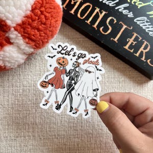 May include: A Halloween-themed sticker featuring three costumed figures: a pumpkin-headed character, a skeleton, and a ghost, each holding a jack-o'-lantern. The sticker has the text "Let's go ghouls" and is decorated with bats. A black sign with the word "MONSTER" is in the background.