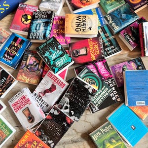 May include: A collection of miniature books with colorful covers, featuring titles like "Harry Potter," "A Court of Silver Flames," and "Hunting Adeline." The books are scattered on a wooden surface, showcasing various genres and authors.