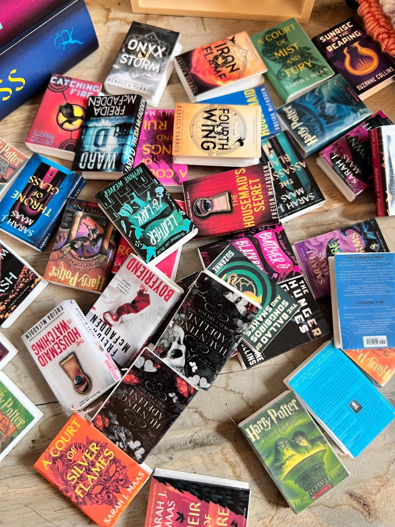 May include: A collection of miniature books with colorful covers, featuring titles like "Harry Potter," "A Court of Silver Flames," and "Hunting Adeline." The books are scattered on a wooden surface, showcasing various genres and authors.