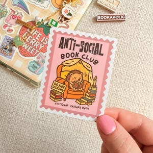 May include: A pink sticker featuring the words "ANTI-SOCIAL BOOK CLUB" and a cartoon hedgehog reading a book. The sticker also includes the text "DO NOT DISTURB" and "FICTIONAL FRIENDS ONLY". Other stickers and accessories are visible.