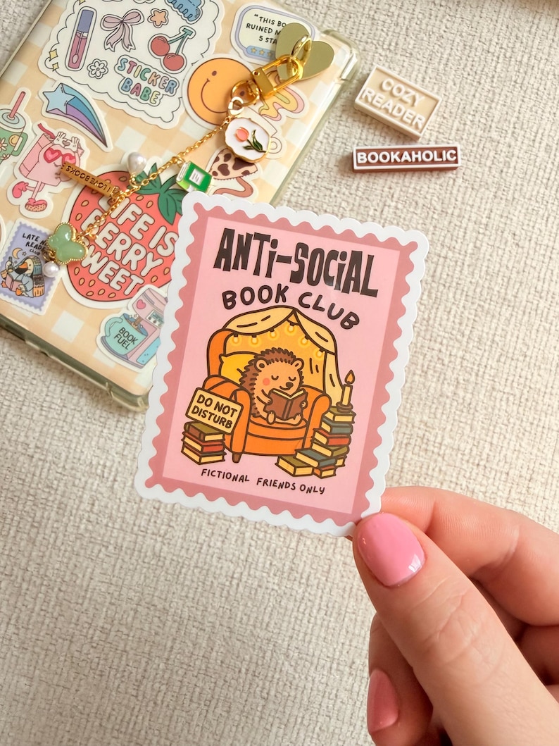 May include: A pink sticker featuring the words "ANTI-SOCIAL BOOK CLUB" and a cartoon hedgehog reading a book. The sticker also includes the text "DO NOT DISTURB" and "FICTIONAL FRIENDS ONLY". Other stickers and accessories are visible.