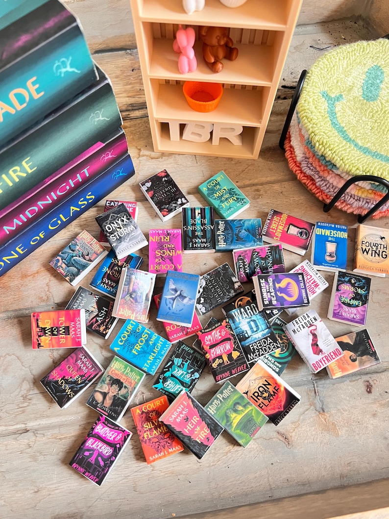 May include: A collection of miniature books with colorful covers, scattered on a wooden surface. Titles include "Harry Potter," "A Court of Thorns and Roses," and "Housemaid." A small wooden bookshelf and a stack of smiley face coasters are also visible.