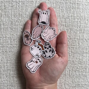 Peut inclure: Une collection de dix autocollants fant&ocirc;mes dans diff&eacute;rentes poses, dont un fant&ocirc;me portant un chapeau de cowboy et un fant&ocirc;me avec un bouquet de fleurs. Les autocollants sont sur une main, et ils sont tous blancs avec des contours noirs.