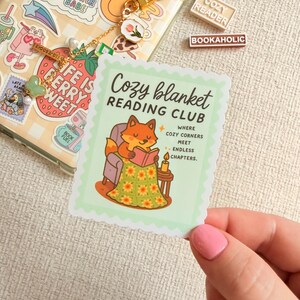 May include: A sticker featuring a cartoon fox reading under a blanket, part of the "Cozy Blanket Reading Club." The sticker has a light green scalloped edge. Other stickers and accessories are visible in the background.