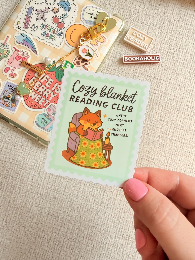 May include: A sticker featuring a cartoon fox reading under a blanket, part of the "Cozy Blanket Reading Club." The sticker has a light green scalloped edge. Other stickers and accessories are visible in the background.