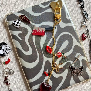 May include: A clear phone case with a black and white zebra print design. The case is adorned with various charms, including a gold heart clasp, a red heart, a rose, and a skull butterfly. Additional charms are visible on a separate chain.