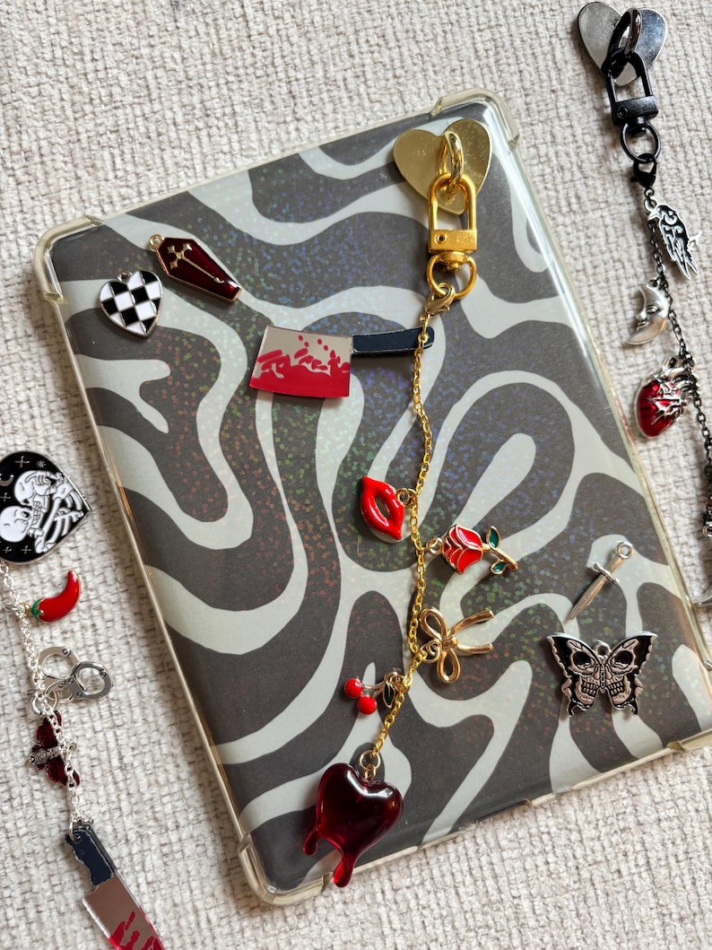 May include: A clear phone case with a black and white zebra print design. The case is adorned with various charms, including a gold heart clasp, a red heart, a rose, and a skull butterfly. Additional charms are visible on a separate chain.
