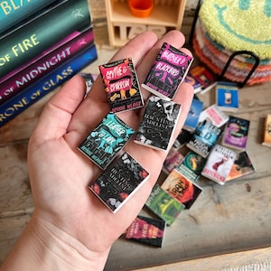 May include: Miniature book collection featuring titles like "Scythe & Sparrow" and "Butcher & Blackbird." The small books, with colorful covers, are held in a hand, with a stack of larger books in the background. A small wooden shelf and a smiley face coaster are also visible.