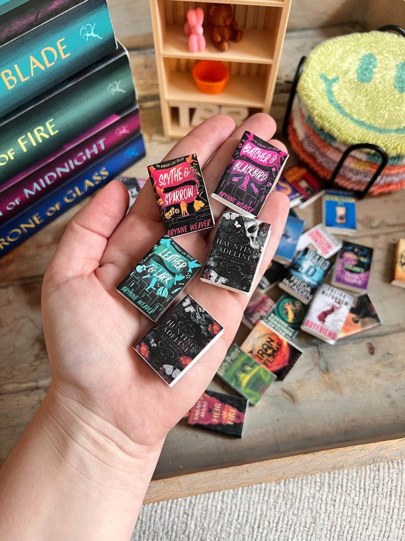 May include: Miniature book collection featuring titles like "Scythe & Sparrow" and "Butcher & Blackbird." The small books, with colorful covers, are held in a hand, with a stack of larger books in the background. A small wooden shelf and a smiley face coaster are also visible.
