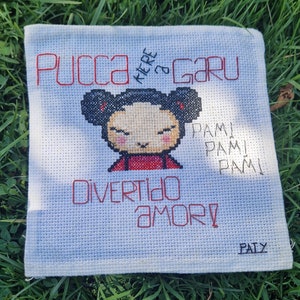 May include: A white cross-stitch embroidery with a cartoon character, Pucca, and the text "Pucca KERE GARU PAM! PAM! PAM! DIVERTIDO AMOR! PATY".